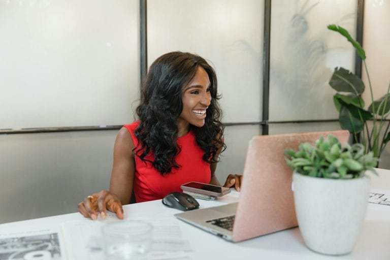 Professional black woman smiling at desk using laptop and smartphone in office.